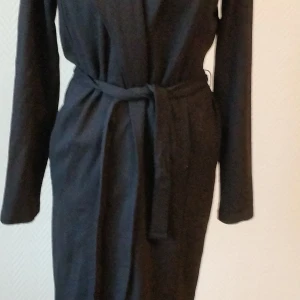 City coat från MeandI - Använd EN gång, men var "inte jag" Lång svsrt kappa i kraftig trikå som ger ett nästan vävt utseende. Fickor på vardera sida och en knapp fram. Bälte att knyta i midjan. Längden slutar vid knäet. 70% viscose,  25% polyester, 5% elastan SMALL men passar även M