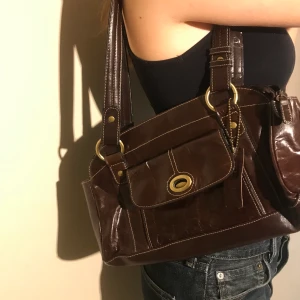 Väska 2000-tal - Brun, glansig läderväska köpt på secondhand. Tre stycken fack och väldigt rymlig. Helt oanvänd och fint skick!🥰köparen står för frakt!                            vid stort intresse vinner högst budande, (buda i kommentarsfältet)annars är det först till kvarn☺️ väskans HÖJDxBREDD: 18x29cm. Handtagets längd (hela bågen): 59 cm.                               