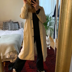 Monki Trenchcoat - En ljus trenchcoat med fin passform som går att stänga med knappar och band i midjan. Klassisk modell. En liten fläck vid armbågen (se bild tre), syns på nära håll. Säljer pga för liten. Tar emot betalning via swish och köparen står för frakten