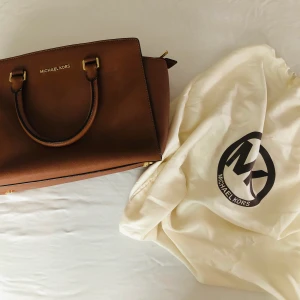 Michael Kors Selma Satchel Tan - Nypris: 3695. Säljs för 2000. Nyskick.  Väskan har dubbla handtag upptill samt en axelrem i äkta läder som kan justeras.  - Försluts med dragkedja - Väskan har fyra innerfack varav ett försluts med dragkedja - Dustbag medföljer -Längd 34 cm, bredd 10 cm, höjd 21 cm.