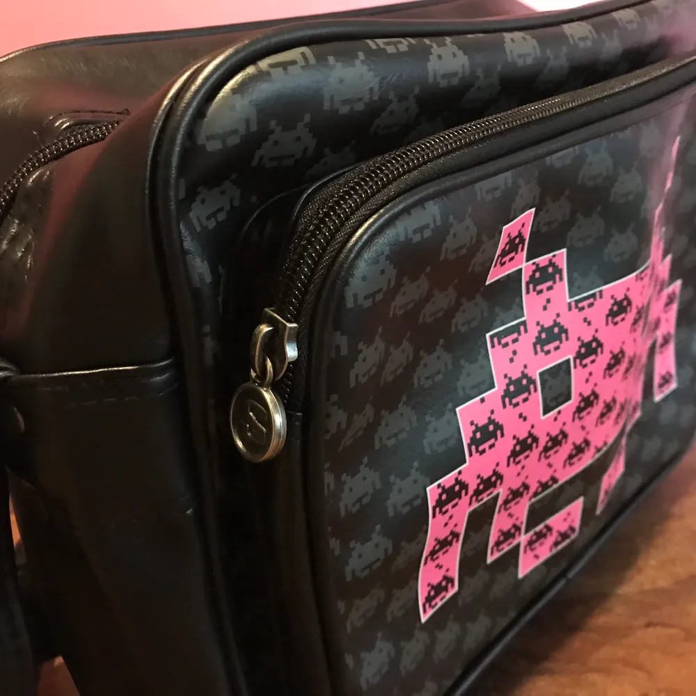 Helt ny väska med retro spelet Space Invaders motiv. Svart väska med rosa motiv i storlek 30*24*10cm. Märket på väskan är SkyLine. Axelremsväska som kan hämtas upp i Enskededalen eller skickas mot fraktkostnad.. Väskor.