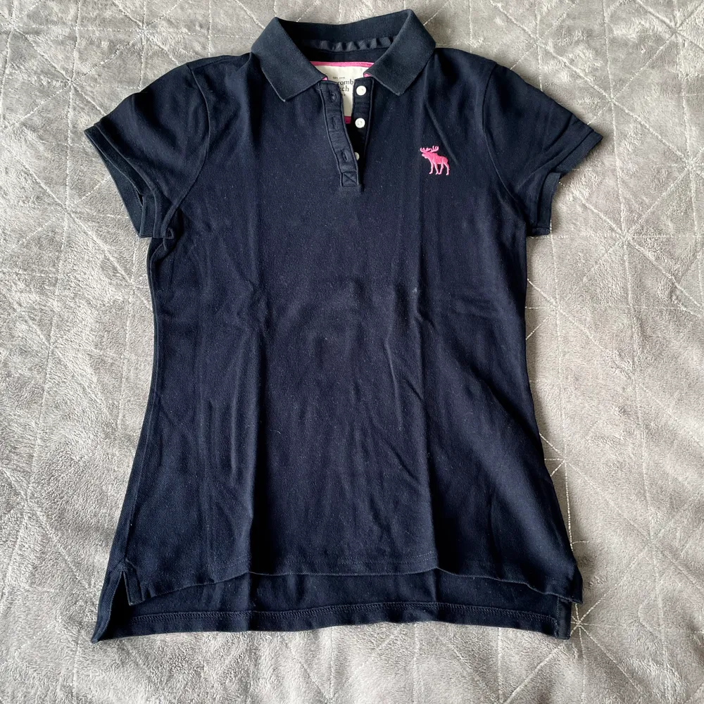 En mörkblå Abercrombie piké med rosa detaljer. I fint skick. Den är storlek M men är liten i storleken.. T-shirts.