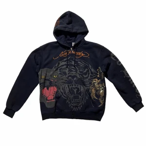 Svart Ed Hardy hoodie - Skitfet svart Ed Hardy zip-up hoodie med tryck och kristaller. Toppskick, inga defekter. Köp direkt för 700kr. BUD ÄR BINDANDE!