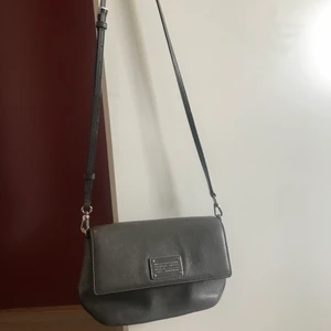 Marc Jacobs väska - Säljer en liten grå Marc Jacobs väska med justerbart axelband tillsammans med matchande kort/nyckelhållare. Båda två har silverdetaljer och är endast använda 1 gång så dem är i väldigt fint skick. Inköpta för många år sedan och finns inte att köpa idag. Väskan har fack för kort samt ett fack med dragkedja. Köparen står för frakten.
