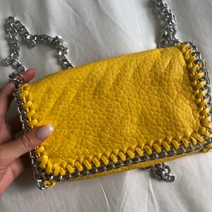 Snygg axelremsväska  - Snygg gul axelremsväska i skinnimitation kantad med silverfärgad kedja. Ett fack med dragkedja på insidan. Den har en avtagbar axellänk som är 105 cm lång. Väskans längd 21 cm, höjd 14 cm, bredd 5cm. Väskan är i nyskick, perfekt julklapp!🥰