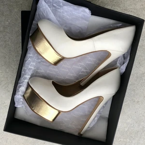 Snygga vita eleganta högklackade skor. - Snygga vita eleganta högklackade skor med gulddetaljer.   Märke: NLY.  Storlek: 36 Högklackade skor, klacken ca 14,5cm och platån är på 3,5-4cm. ( gulddetaljen)   Pris: 70 kr + frakt 66 ( spårbart)  Skick : Dem är sparsamt använda, har inget direkt att anmärka förutom 1 repa framtill och lätt skavd vid innerkanterna, samt mini rispa vid guldet.  Det är skolim som syns i skon, finns även synligt vid ena klacken.  Se bilder.  Skorna kommer från rökfrit och djurfritt hem.