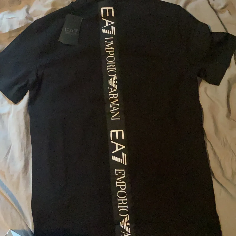 En svart Emporio Armani t-shirt med svart ”EA7 Emporio Armani” logo på bröstet samt vit text på ryggen. Tröjan är aldrig använd och kommer tillsammans med påsen, köpt på NK. Anledning till varför jag säljer är att den ej passade mig storleksmässigt. Urpringspris: 1000kr. T-shirts.