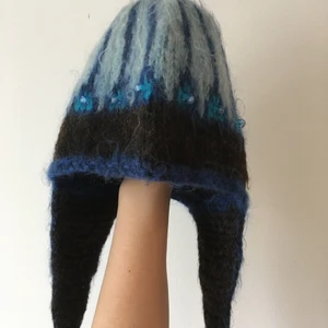 Handstickad, blå ullmössa med öronlappar - Superfin handstickad mössa från Kiruna. Pärlor instickande runt hela mössan och en rund, svart knapp att knäppa ihop öronlapparna under hakan om det är riktigt kallt. Den är i ull och jättevarm och skön. Lite 1970-tals känsla.