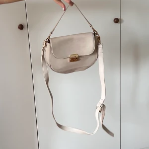 beige väska - Beige väska (skinnimitation) från NAKD med gulddetaljer. Går att ha den som handväska eller axelremsväska och den är justerbar. Använd men bra skick. Hör av dig om du undrar något🤍