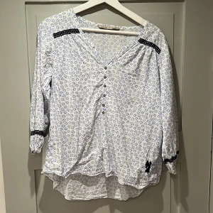 Mönstrad House of Lola-blus - Vit blus med blåblommigt mönster och detaljer såsom knappar, broderiband och Lola-ängeln. Fin till sommaren, luftig och skön bomull. 