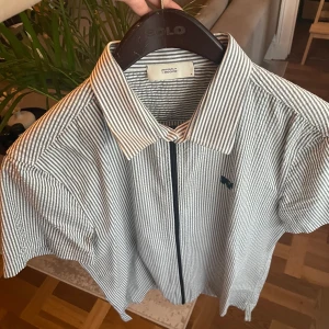 Uniforms for the dedicated kortärmad skjorta - Kortärmad kritstrecksrandig skjorta i crinkled bomull. Med svart dragkedja och märkets logga embroderad på bröstet. Väldigt unik och otrolig bra kvalitet skjorta köpt på NK för ca 1200kr