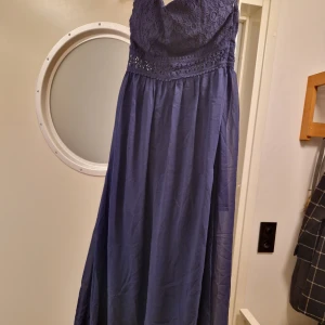 Mörkblå balklänning  - Lång mörkblå ärmlös festklänning säljes. Fin spetsdekoration över bysten och ryggen.  Överdelen är elastisk och stängs med dold dragkedja i sidan. Silikonband på övre främre insida. Nederdelen har ett snyggt flowy-fall. Inköpt i NY 2013 använd 1 gång