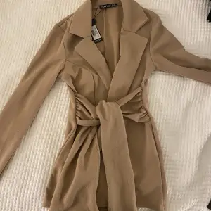 En figursydd blazer i beige/khaki färg. Aldrig använd då den var för liten, lappen sitter kvar. Den är från boohoo. Storlek 34. Tredje bilden är från modellen på hemsidan. 