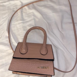 Väska - Säljer denna beige/ljusrosa väskan med gulddetaljer. Har användt den ett par gånger så tyget inne har lossnat lite, men det kan lätt limmas på igen. Bandet kan gå av så att det blir en liten fin handväska. Pris kan sänkas vid snabb affär.