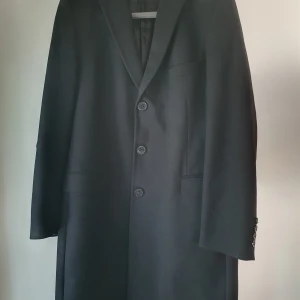 Svart ullrock strl 54 - Tidlös herrock från David Design. Mycket fin! Enda anmärkning lite slitet vid innerficka.  Mått i cm: Midjevidd 112 Axel till axel 51 Ärmlängd 70 Ryggbredd där sömmar går ihop i höjd med skuldror 48 Ärmhåla till ärmhåla 60 Rygglängd 103 Bredd nederkant 66