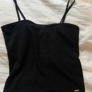 Svart linne med mönster - Ett svart kortare linne med fint mönster. Rätt smala axelband och en rak passform. Hel och fin bara blivit lite kort då materialet inte är stretchigt utan den sätter sig rätt tajt! 