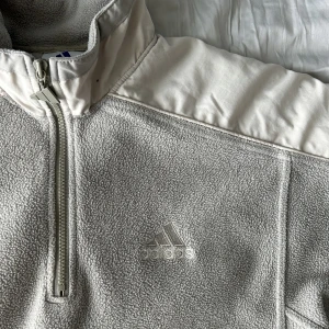 Vintage adidas fleece - Vintage-oversized adidas fleece i bra skick. Tröjan är grå med mintgröna toner. Passformen går att justera genom att dra åt ett snöre i tröjan.