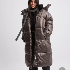 Oanvänd lång dunjacka strl. 34 (oversized) från NA-KD - Brun-aktig glansig dunjacka i storlek 34 från NA-KD. Säljer pga missade returdatum och den var för stor för mig (oversized), passar snarare en 36-38. Modellen är så najs och färgen är jättefin!  Helt oanvänd, lapp sitter kvar. Nypris: 1199kr