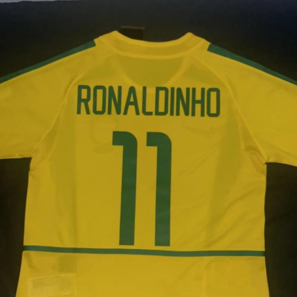 Fet retro Brasilien fotbollströja med ikonen Ronaldinho 11 på ryggen🇧🇷. Säljer pågrund av liten storlek. Perfekt till sommaren. Tröjan är helt ny med etikett kvar. Fick den i present. Skriv vid funderingar.. Sport & träning.