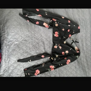 Svart blommig omlottopp från Divided - En svart omlottopp från HM, Divided med blommigt mönster i rosa och grönt. Toppen har långa ärmar och knytning i ryggen.