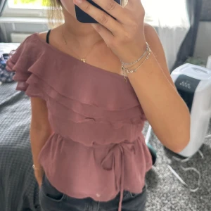Rosa offshoulder topp med volanger - En supersöt rosa offshoulder topp med volanger. Den har ett knytband i midjan för att justera passformen. Säljer den då den inte kommer till användning💕💕