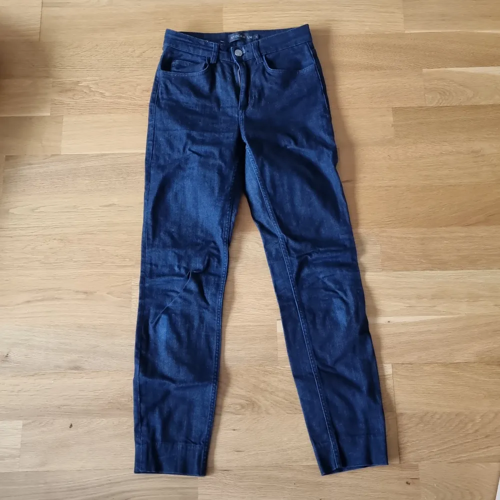 Ett par mörkblå jeansbyxor från Stockh LM i storlek 34. Byxorna är tillverkade av 90% bomull, 6% polyester och 2% elastan, vilket ger dem en bekväm och stretchig passform. De har en klassisk design med fem fickor och knappstängning.. Jeans & Byxor.