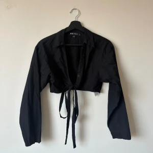 Cropped svart skjorta från Morel Rocks - En kort skjorta med knytning! Man kan knyta banden runt kroppen och göra en rosett/knytning både fram och bak. Supercool och lite annorlunda. Mycket sparsamt använd så är typ i ny skick!