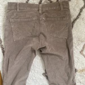 Beige manchesterbyxor från Here+There - Ett par beige manchesterbyxor från märket Here+There. Byxorna har en klassisk femficksdesign med bälteshällor och knappstängning. De är tillverkade i ett mjukt och bekvämt manchestertyg.