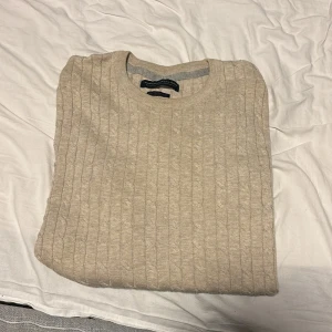 Beige stickad tröja från Jack & Jones - En beige stickad tröja från Jack & Jones med ett kabelstickat mönster. Tröjan har långa ärmar och ribbstickade muddar vid ärmslut och nederkant.