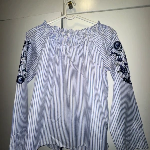 Randig offshoulder blus med broderi - En jättefin randig offshoulder blus med blå och vita ränder. Blusen har långa ärmar och broderade blommor på ärmarna💗💗