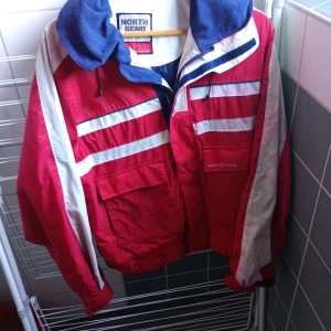 Röd och vit vindjacka från North Bend - En röd och vit vindjacka från North Bend med dragkedja och fickor. Jackan har en blå insida och en gul huva. Den har långa ärmar och är lämplig för vår- och höstsäsonger.