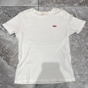 Vit t-shirt från Levi's - En klassisk vit t-shirt från Levi's med korta ärmar och en liten röd Levi's-logga på bröstet. T-shirten är tillverkad i bomull och har en normal passform.