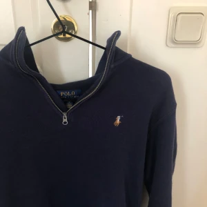 Mörkblå tröja från Ralph Lauren - En mörkblå tröja från Ralph Lauren med dragkedja vid halsen och en broderad logotyp på bröstet. Tröjan har långa ärmar och är tillverkad i ett bekvämt material. Passar dig som bäst 170-175