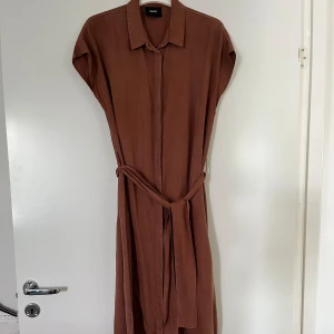 Skjortklänning Object 38 - Brun skjortklänning från Object, storlek 38. Stängs med knappar, se bild. Skärp i midjan som går att ta av. Små snitt på sidorna. Klippt av lapp på material, misstänker Lyocell. Klänningen är i mycket god skick.   Mått: Längd ca 126 cm Byst ca 52 cm 