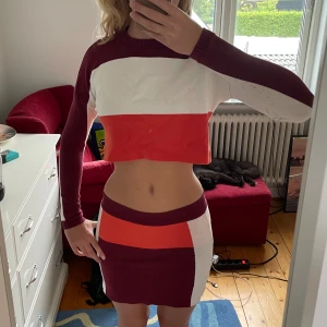 Flerfärgad croppad tröja och kjol från Urban Outfitters - Säljer en supercool set från Urban Outfitters i storlek XS. Setet består av en croppad tröja och en tight kjol. Båda plaggen har ett färgglatt mönster i vinröd, vit och orange. Tröjan har långa ärmar och är ribbad vid ärmslut och hals. Materialet känns mjukt och skönt mot huden.