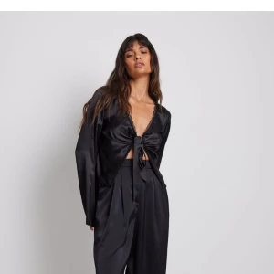 Svart satinblus med knytning - Säljer en elegant svart blus i satinmaterial. Blusen har långa ärmar och en knytning framtill som ger en snygg detalj. Perfekt för festliga tillfällen eller en utekväll. Den har en djup v-ringning med spetsdetaljer längs kanterna. Passar bra till både byxor och kjol för en chic look.