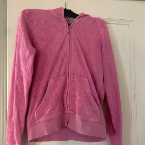Rosa hoodie från Juicy Couture - Säljer en superfin rosa hoodie från Juicy Couture. Den har en dragkedja framtill och en stor ficka där fram. På baksidan finns ett snyggt broderat märke med två svarta hundar, ett hjärta och en krona. Hoodien är ganska mycket använd. Perfekt för både mysiga hemmakvällar och casual outfits. Passa på att fynda denna fina  hoodie💗