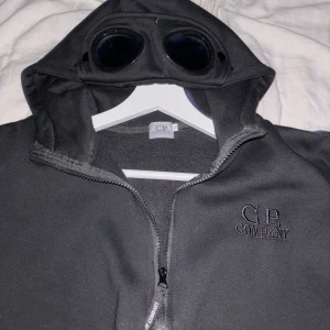Svart jacka från C.P. Company - Säljer en svart jacka från C.P. Company. Jackan har en unik design med inbyggda glasögon i huvan och en dragkedja framtill. Perfekt för en cool och annorlunda look!
