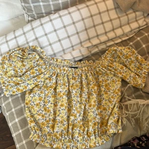 Gul blommig off-shoulder topp - Säljer en superfin gul off-shoulder topp med blommigt mönster. Toppen har korta ärmar och en elastisk kant vid axlarna och midjan för en bekväm passform. Perfekt för sommaren och festliga tillfällen! 🌼