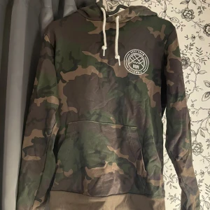 Camouflage hoodie från Dope - Säljer en snygg camouflage hoodie från Dope. Den har en stor ficka på magen och en vit logga på bröstet och ryggen. Perfekt för höst och vinter, och den är i nyskick. Den är gjord av bomull och polyester, vilket gör den både bekväm och hållbar. Passformen är normal och den har en justerbar huva med vita snören.
