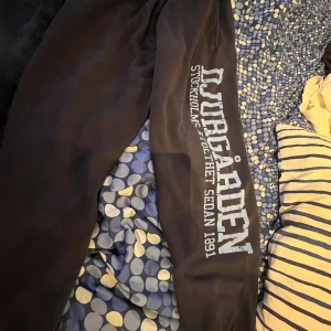 Svarta mjukisbyxor från Djurgården - Säljer ett par svarta mjukisbyxor från Djurgården. Byxorna har texten 'Djurgården Stockholm Stolthet Sedan 1891' tryckt längs benet. Perfekta för en avslappnad dag hemma eller för att visa ditt stöd för laget. De är bekväma och har en normal passform.