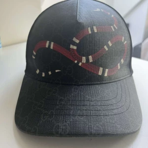 Svart keps från Gucci med ormmönster - Säljer en snygg svart keps från Gucci med ett coolt ormmönster i rött och vitt på framsidan. Kepsen är i storlek XS och har en justerbar rem baktill för perfekt passform. Materialet är av hög kvalitet och kepsen är i nyskick. Perfekt för att ge din outfit en lyxig touch!