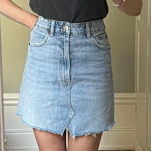Ljusblå jeanskjol med rå kant - Superfin ljusblå jeanskjol med rå kant. Kjolen har en klassisk femficksdesign, sliten look och en liten slits framtill. Lätt att styla både till vardag och fest! Midjemått tvärs över: 33 cm. Längd: 40 cm. 
