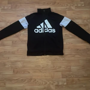 Svart Adidas träningsjacka med vita detaljer - Säljer en svart Adidas träningsjacka i storlek S. Jackan har vita detaljer på ärmarna och en stor vit Adidas-logga på framsidan. Den är gjord av polyester och har en dragkedja framtill. Perfekt för träning eller vardagsbruk!