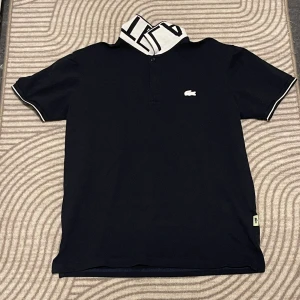 Svart pikétröja från Lacoste - Säljer en snygg svart pikétröja från Lacoste. Den har en cool vit krage med svart text och en liten broderad krokodil på bröstet. Tröjan är i bra skick och perfekt för både vardag och fest. Passa på att köpa denna stiliga tröja! 🐊