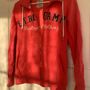 Zipup - Korallröd Zipup från Etirel i storlek XS, men sitter mer som S. Mörkblått och vitt ”tryck”. Välanvänd men har inga fläckar eller hål. Texten på tröjan är dock lite nopprig.