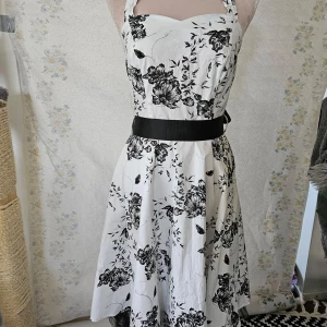 Vit och svart blommig klänning - En elegant 50-tals inspirerad vit klänning med svart blommönster. Klänningen har en svart knytbälte i midjan och är axelbandslös med en dragkedja på baksidan. Materialet består av 65% bomull och 35% polyester.