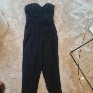 Svart axelbandslös klänning - En elegant svart axelbandslös byxdress i medium storlek. Klänningen har en figurnära passform upptill och en något lösare passform nedtill. Perfekt för festliga tillfällen.