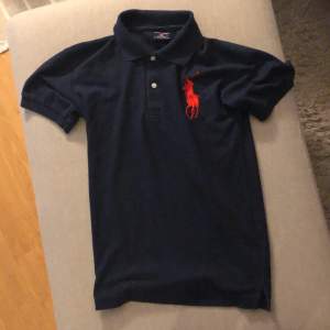 En mörkblå pikétröja från Polo Sport Ralph Lauren i storlek M. Tröjan är tillverkad av 100% bomull och har en röd broderad logotyp på bröstet. Den har korta ärmar och en klassisk krage med två knappar. Den är helt i nyskick!