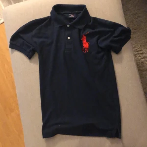 Mörkblå pikétröja från Polo Sport Ralph Lauren - En mörkblå pikétröja från Polo Sport Ralph Lauren i storlek M. Tröjan är tillverkad av 100% bomull och har en röd broderad logotyp på bröstet. Den har korta ärmar och en klassisk krage med två knappar. Den är helt i nyskick!